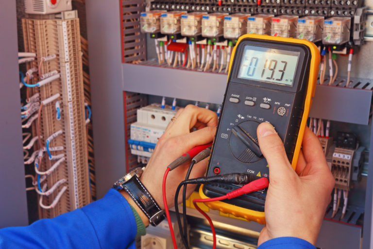 A person holding a multimeter in front of a wall of electrical equipment